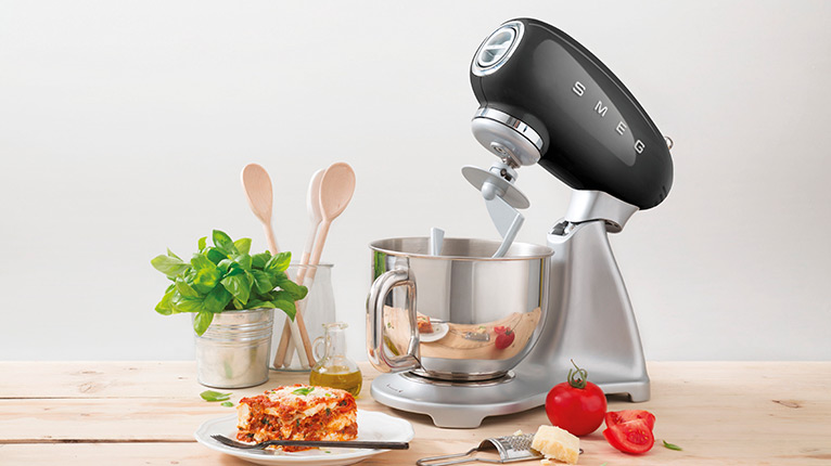 smeg Küchenmaschinen bei Moebel-Shop – Schwarze Küchenmaschine auf heller Holzarbeitsplatte. Umgeben von Kochutensilien, frischen Zutaten und einem angerichteten Teller Lasagne – Genuss trifft Stil.