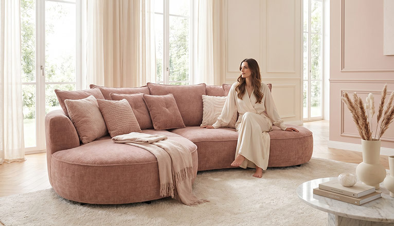 Frau sitzt auf einem rosafarbenen Sofa in einem hellen Wohnzimmer mit großen Fenstern, weichen Textilien und eleganten Wohnaccessoires.