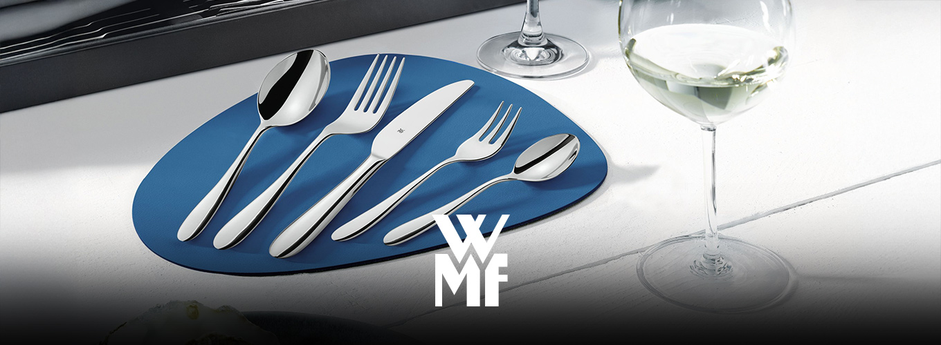 WMF-Besteckset aus fünf Teilen – Suppenlöffel, Gabel, Messer, Kuchengabel und Kaffeelöffel – auf blauer Tischunterlage; daneben steht ein Weinglas.