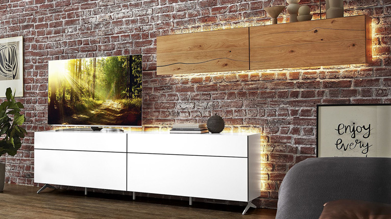 Jetzt ROLF WEBER Wohnen entdecken! - Ein weißes, griffloses ROLF WEBER Lowboard, auf dem ein Fernseher steht und ein beleuchteter ROLF WEBER Hängeschrank aus Holz in einem Wohnzimmer vor einer roten Ziegelwand