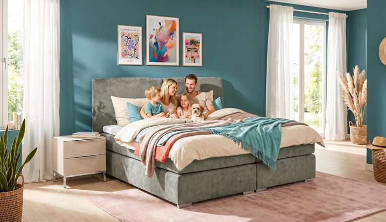 Familie entspannt gemeinsam auf einem Boxspringbett mit Hund in einem hellen Schlafzimmer mit modernen Möbeln und wohnlichen Dekorationen.
