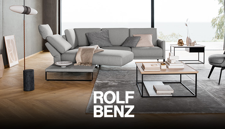 ROLF BENZ Sofas - Ein hellgraues ROLF BENZ Sofa mit schwenkbarem Liegeelement, ein ROLF BENZ Sessel und ROLF BENZ Couchtische in einem Wohnzimmer mit großer Fensterfront und Wänden in Beton-Optik