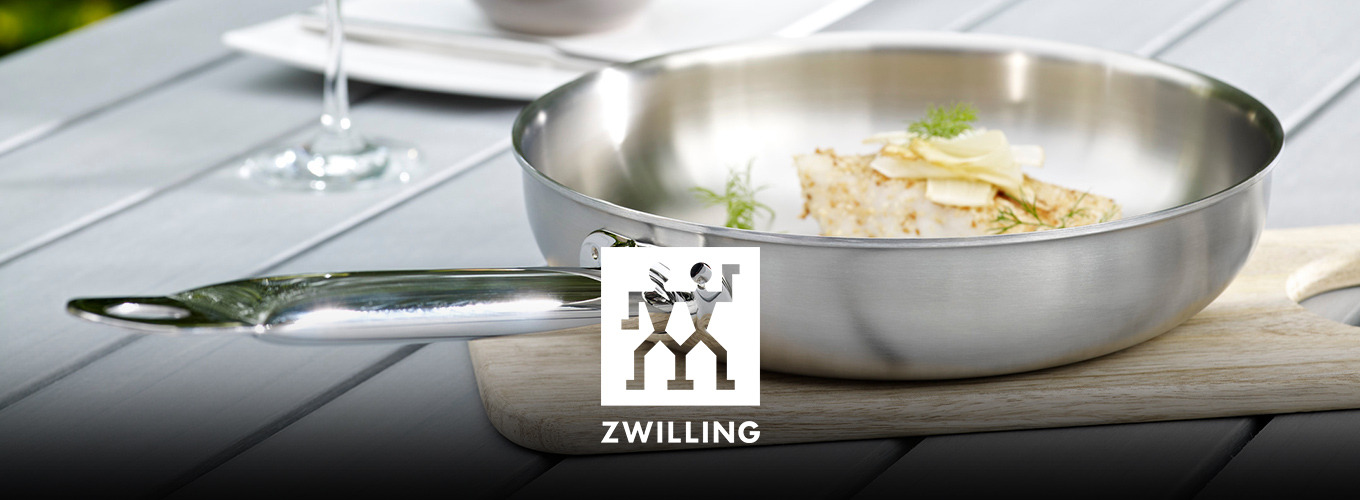 ZWILLING-Edelstahlpfanne mit gekochtem Essen auf einem Holzbrett auf dem Gartentisch; im Hintergrund gedeckter Platz mit Weinglas.